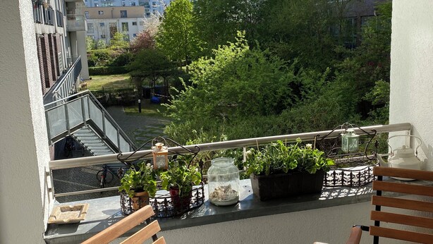 Un balcon avec des chaises et une table en bois, des plantes en pot et une vue sur une rivière et des bâtiments.