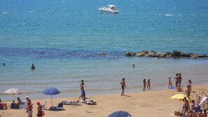 A beach with people sunbathing and swimming, a boat in the water, and umbrellas providing shade. - Apartment with heated saltwater pool, private garden, air conditioning, WiFi (Donnalucata)