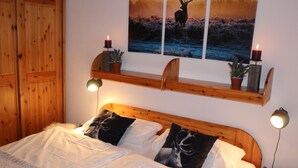 Ett sovrum med en sänggavel i trä, två sänglampor, en säng med vita sängkläder, en hylla med två ljus och en krukväxt samt en tavla med en hjort som hänger på väggen.