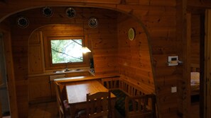 Interior acolhedor de cabana com mesa de jantar de madeira, bancos, janela com vista para árvores e relógio fixado na parede.