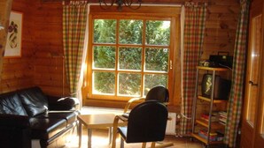 L’intérieur chaleureux d’un chalet, avec un revêtement mural en bois, un canapé en cuir noir, une table à manger en bois, un fauteuil à bascule, un téléviseur posé sur une étagère et une fenêtre agrémentée de rideaux.