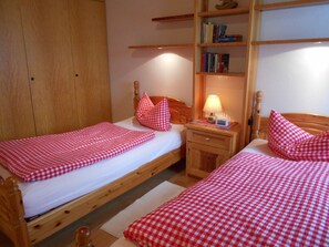 A bedroom with two wooden beds, each with a red and white checkered bedspread and matching pillows. There is a nightstand with a lamp and a book shelf above it.