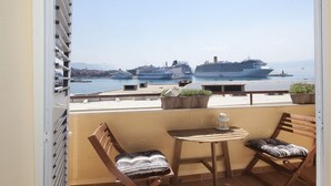 Ein Balkon mit zwei Holzstühlen und einem kleinen Tisch, der auf eine Marina mit mehreren Kreuzfahrtschiffen blickt.