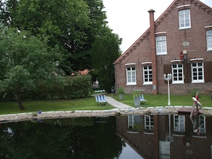 A brick building with a pond in front, surrounded by trees. - Spacious exclusive apartment on the 1st floor with a natural pond (Wittmund-Buttforde)