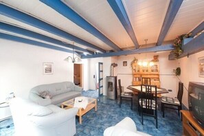 A living room with a sofa, coffee table, and dining table with chairs. There is a fireplace, a television, and a ceiling with exposed beams. - Spacious exclusive apartment on the 1st floor with a natural pond (Wittmund-Buttforde)