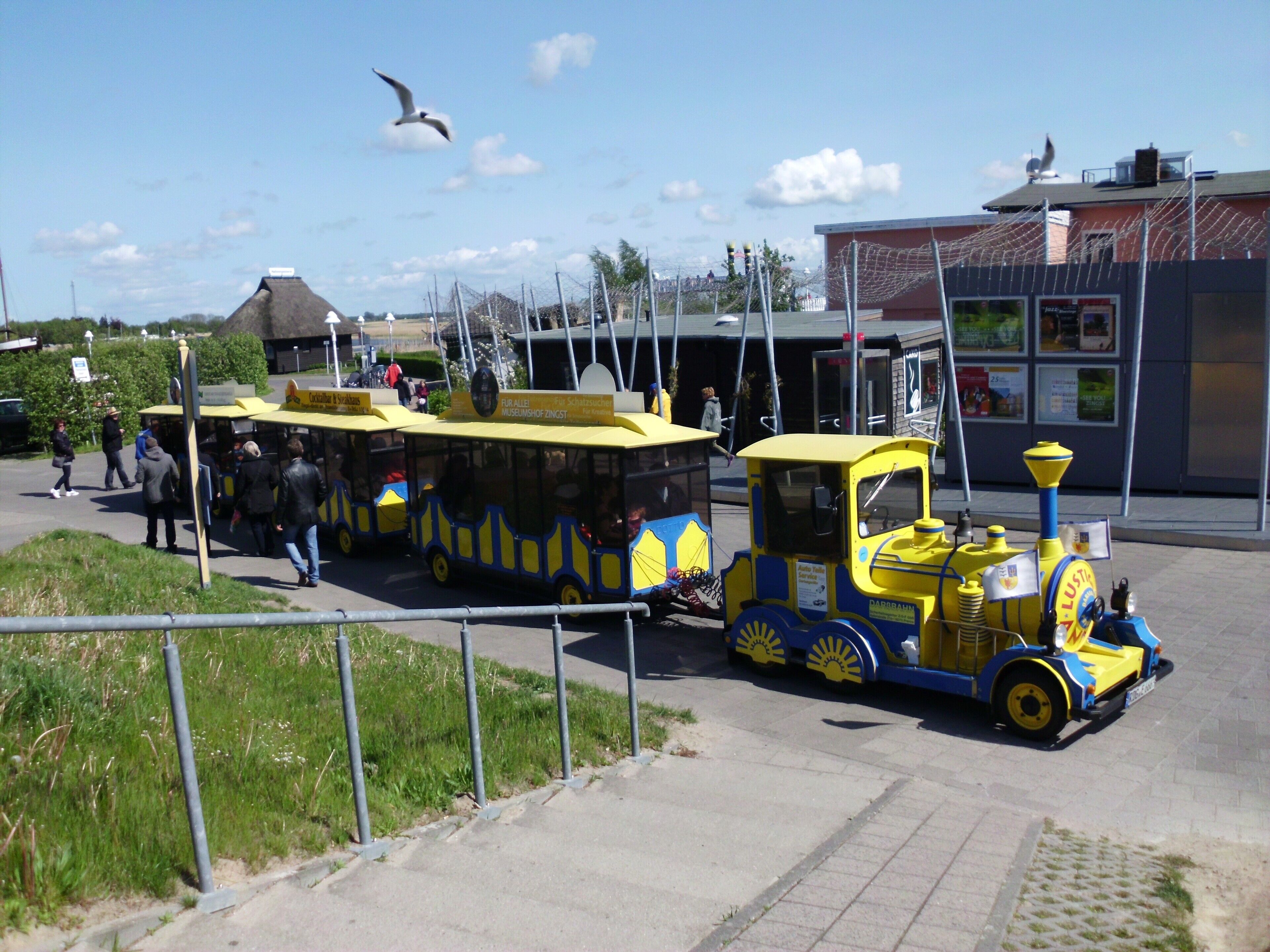 Un train jaune et bleu avec des passagers, un bâtiment avec une enseigne et un ciel dégagé.