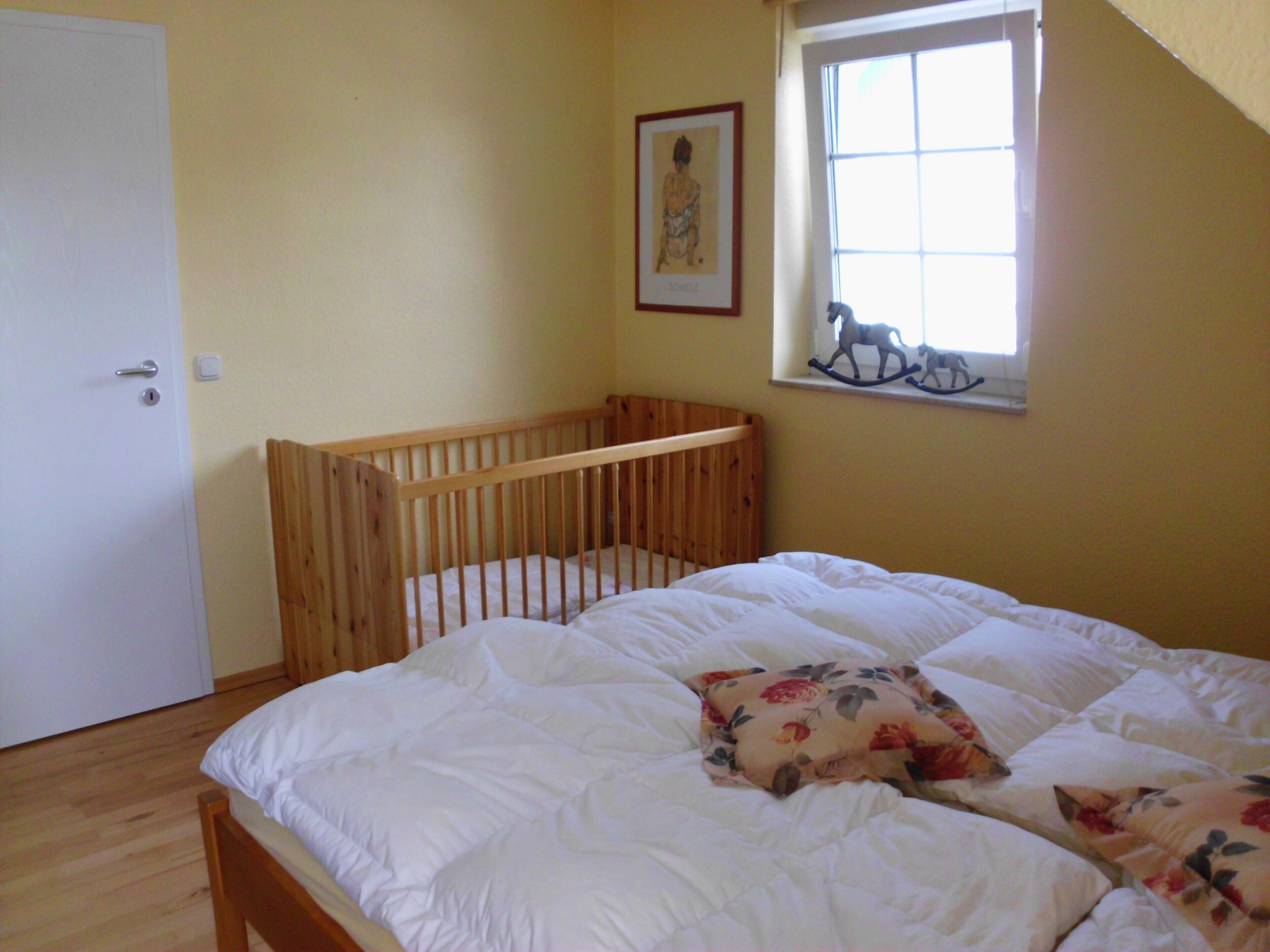 A room with a wooden crib, a bed with floral bedding, a framed picture on the wall, and a window with a rocking horse decoration.