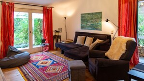 Un salon chaleureux avec un canapé, un fauteuil et un pouf, un tapis coloré et une grande fenêtre ornée de rideaux rouges.