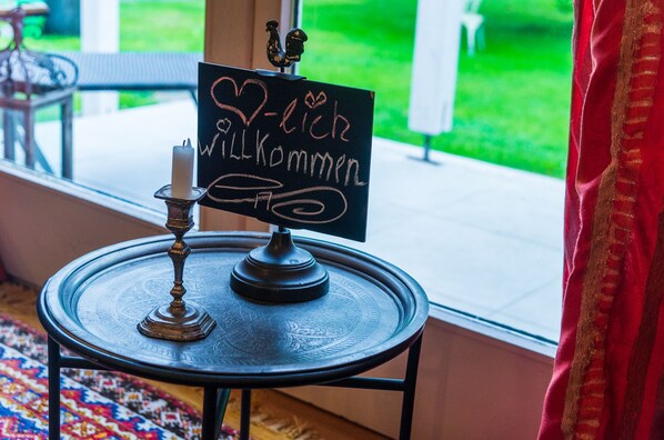 Een schoolbord met 'Willkommen' en een hartsymbool, een kandelaar en een decoratieve vogel op een ronde tafel bij een raam.