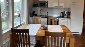 A modern kitchen with white cabinets, a microwave, and a refrigerator. There is a dining table with chairs, and a window with curtains.