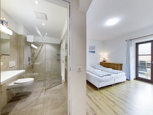 Un baño moderno con cabina de ducha de vidrio, inodoro y lavamanos. El dormitorio tiene una cama con cabecera de madera, una mesita de noche y una ventana con vistas.