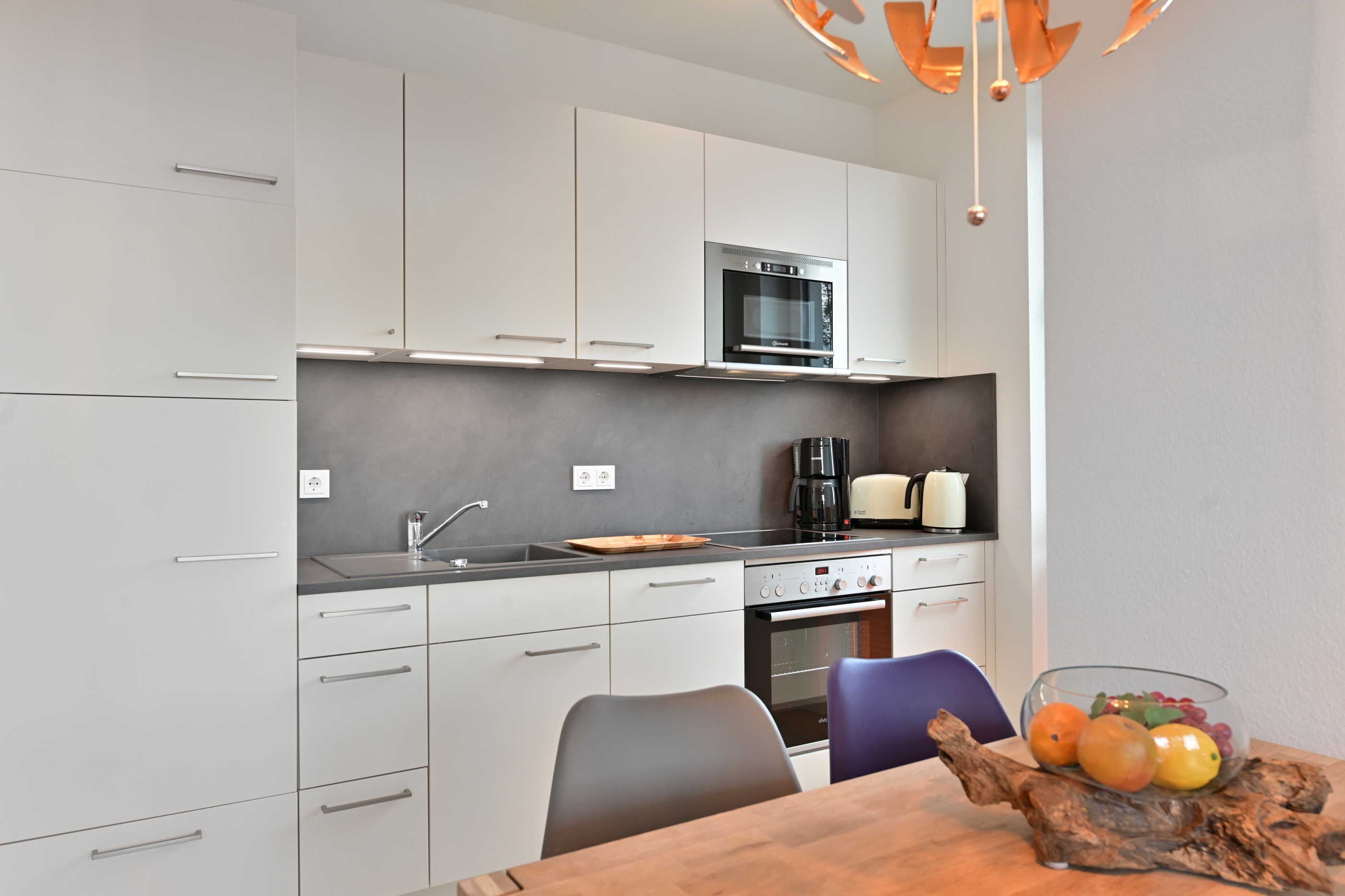 Una cocina moderna con armarios blancos, horno empotrado, cafetera y frutero sobre la mesa.