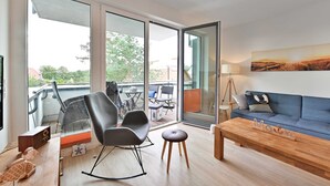 Een moderne woonkamer met een blauwe bank, een houten salontafel en een schommelstoel. Er is een balkon met een glazen deur, een tafel met stoelen en een lamp.