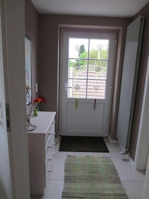 Een smalle gang met een witte deur, een klein vloerkleed en een witte kast met een vaas.