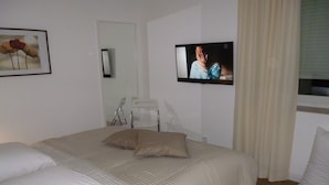 A neatly made bed with white linens, a television mounted on the wall, and a framed picture of flowers. - 5*-Comfort-FeWo, 85m²-new building, 6 pers., HH-South, forest and heath, child-friendly! (Rosengarten-Vahrendorf)