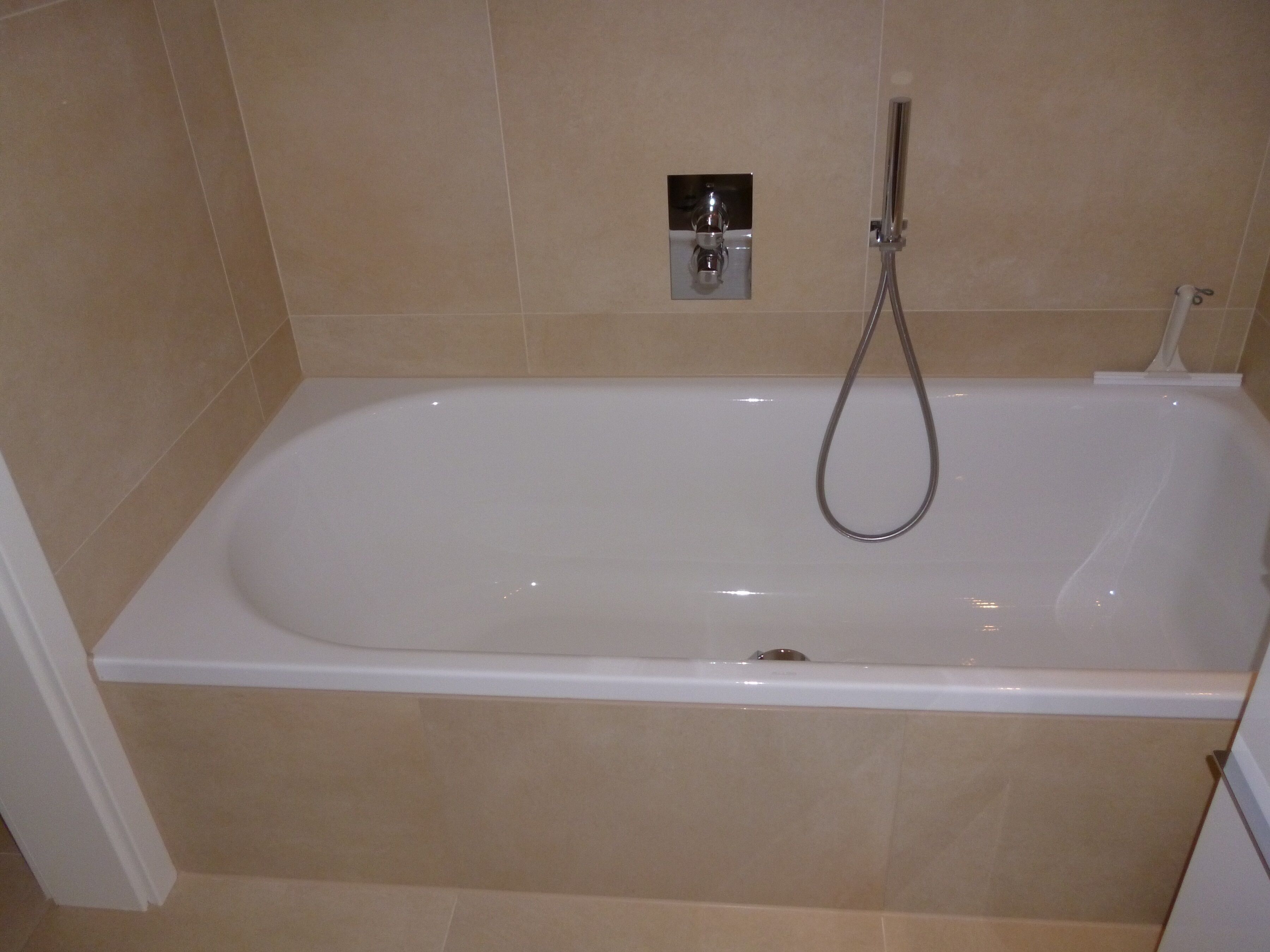 A standard bathtub with a handheld showerhead, beige tiled walls, and a white baseboard.