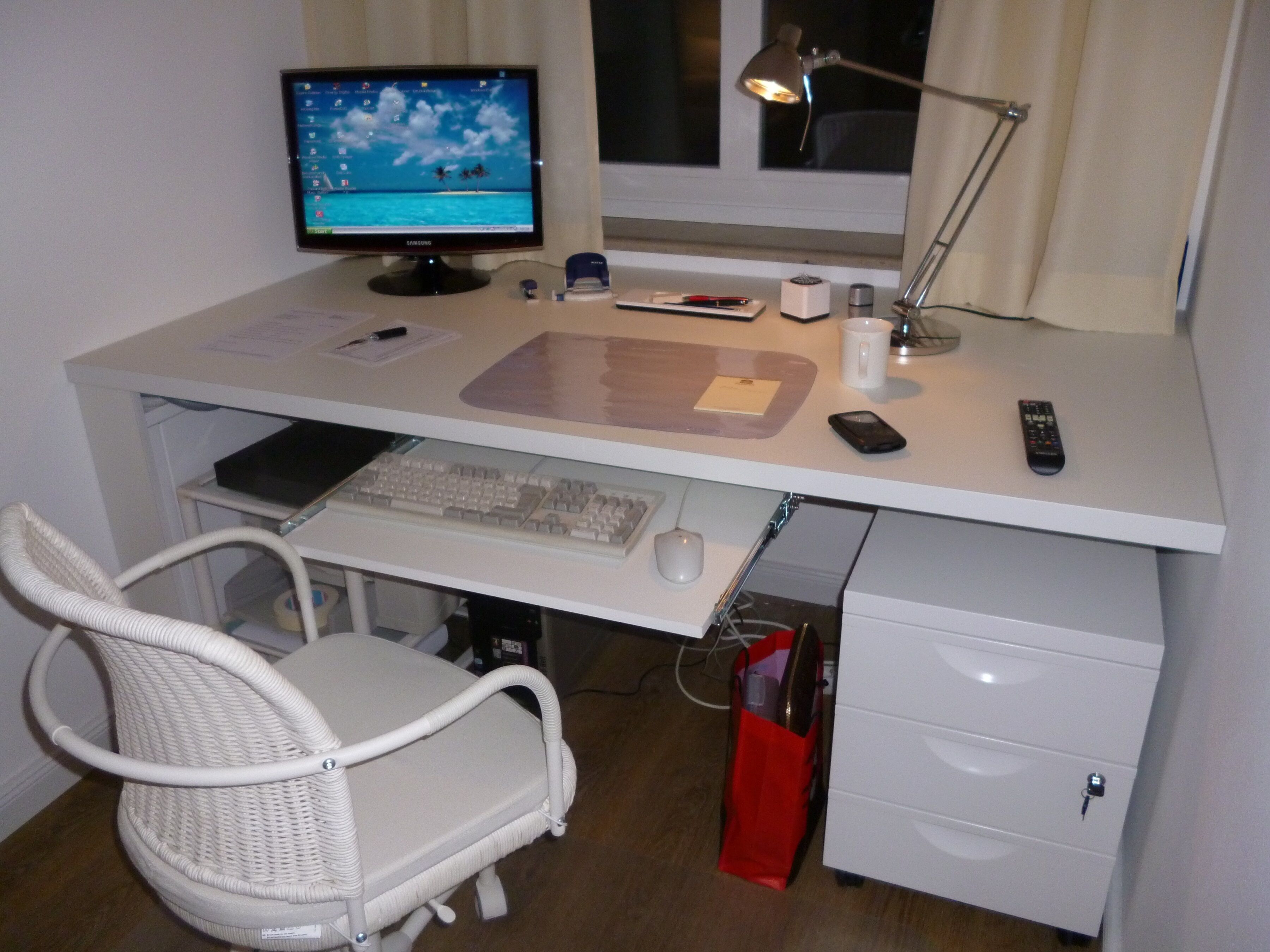 Een wit bureau met een computerscherm, toetsenbord en muis. Er staat een witte stoel, een lamp en een rode tas op het bureau.
