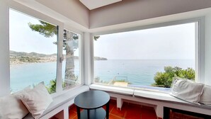 Une pièce d’angle dotée d’une grande baie vitrée donnant sur la côte, d’une petite table ronde et d’un banc agrémenté de coussins.