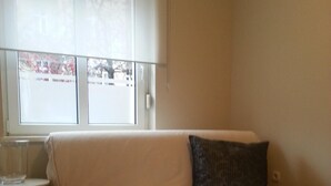 A small, well-lit room with a white sofa, a small table, and a window with a view of a parking lot.