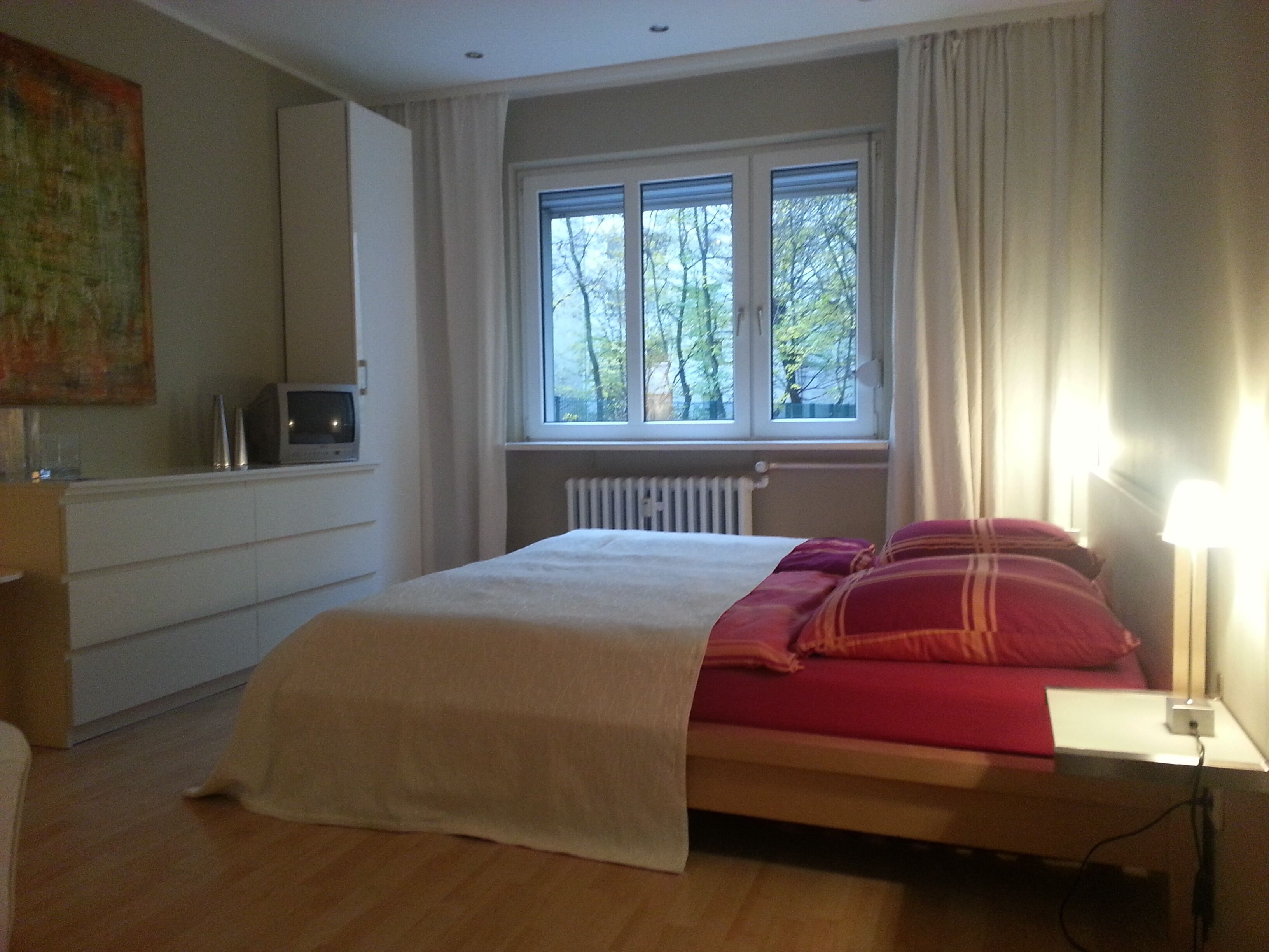 A bedroom with a bed, a window with curtains, a radiator, and a TV on a cabinet.