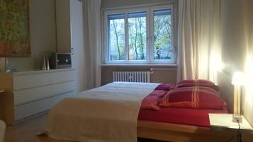 A bedroom with a bed, a window with curtains, a radiator, and a TV on a cabinet.