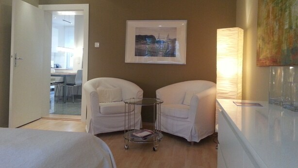 A hotel room with two white armchairs, a small round table, and a bed. There is a painting on the wall and a lamp on the side table.