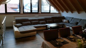 Un salon chaleureux avec un canapé d’angle, une table à manger pour deux personnes et de grandes fenêtres dotées de rideaux.