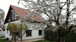 Une maison à deux étages, avec un toit recouvert de tuiles rouges, un petit jardin agrémenté d’un sentier et un arbre aux fleurs blanches.