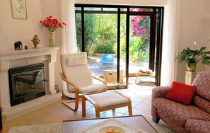 Ein Wohnzimmer mit Kamin, einem weißen Sessel, einem roten, gemusterten Sofa und Blick auf den Garten durch große Glastüren.
