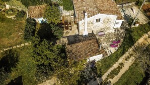 Vue aérienne d’une maison en pierre avec un toit de tuiles, entourée de verdure et bordée d’un chemin de terre.