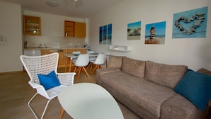 Ein modernes Wohnzimmer mit einer Couch, Sesseln und einem Esstisch. Im Hintergrund sind Küchenschränke und ein Kühlschrank zu sehen.