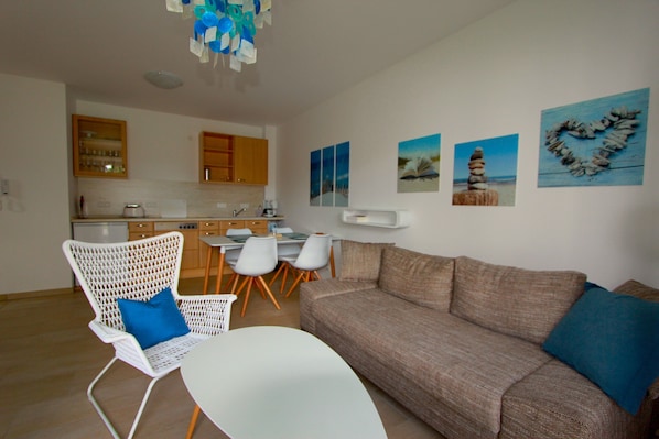 A modern living room with a sofa, chairs, and a dining table. There are kitchen cabinets and a refrigerator in the background.