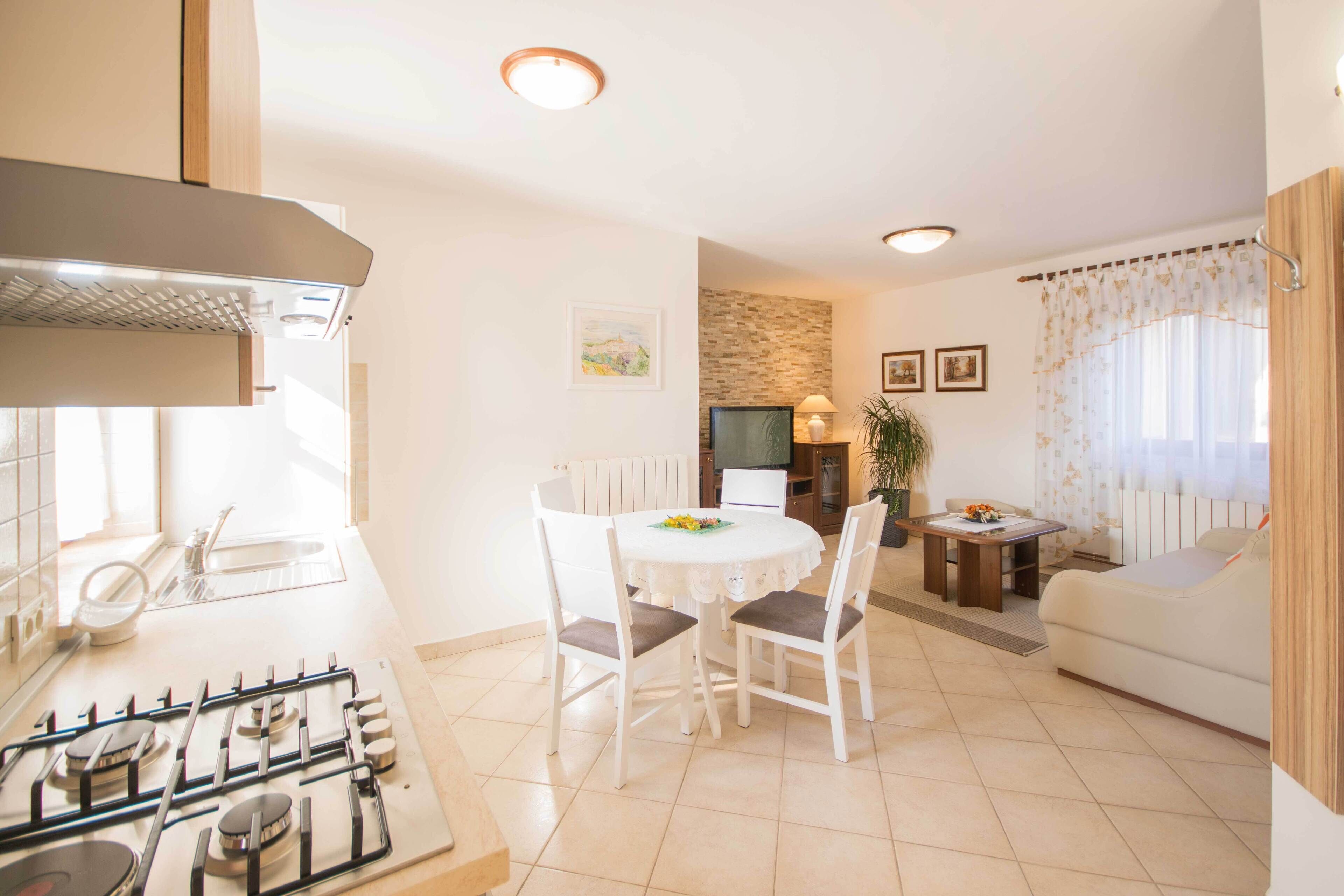 A modern kitchen with a gas stove, white chairs around a dining table, and a living area with a sofa and a TV.