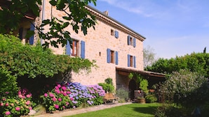 A stone building with blue shutters, a well-maintained lawn, and a variety of flowering plants. - Special prices in autumn! Charming apartment with large, heated pool (Rosières)