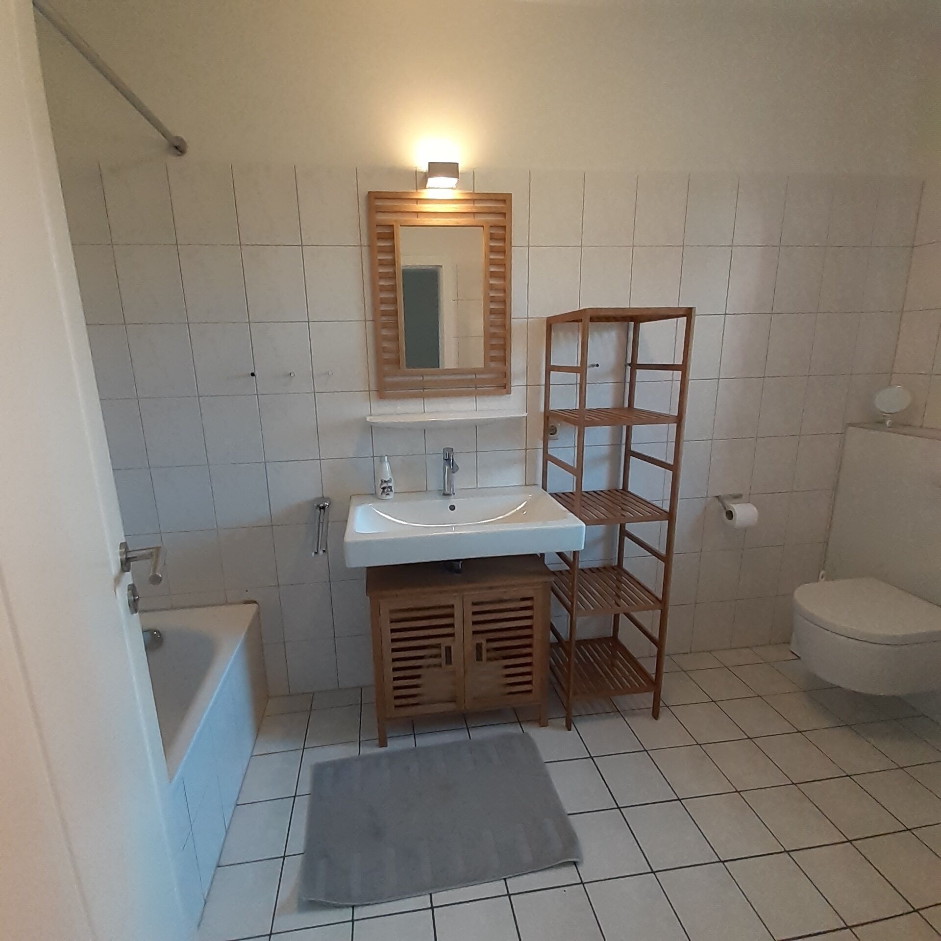 A bathroom with a white sink, mirror, and toilet. There is a wooden shelf unit next to the sink and a small rug on the floor.