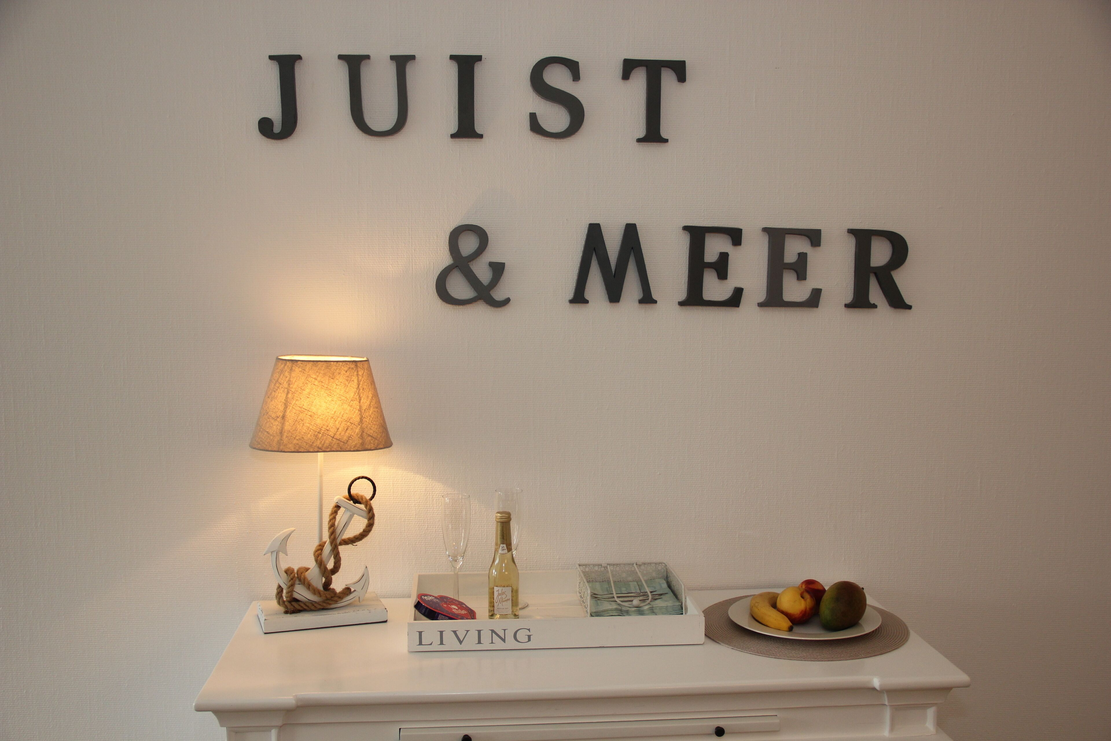Eine Wand mit der Aufschrift 'JUIST & MEER', ein kleiner Tisch mit einer Lampe, einer Flasche und einer Fruchtschale.