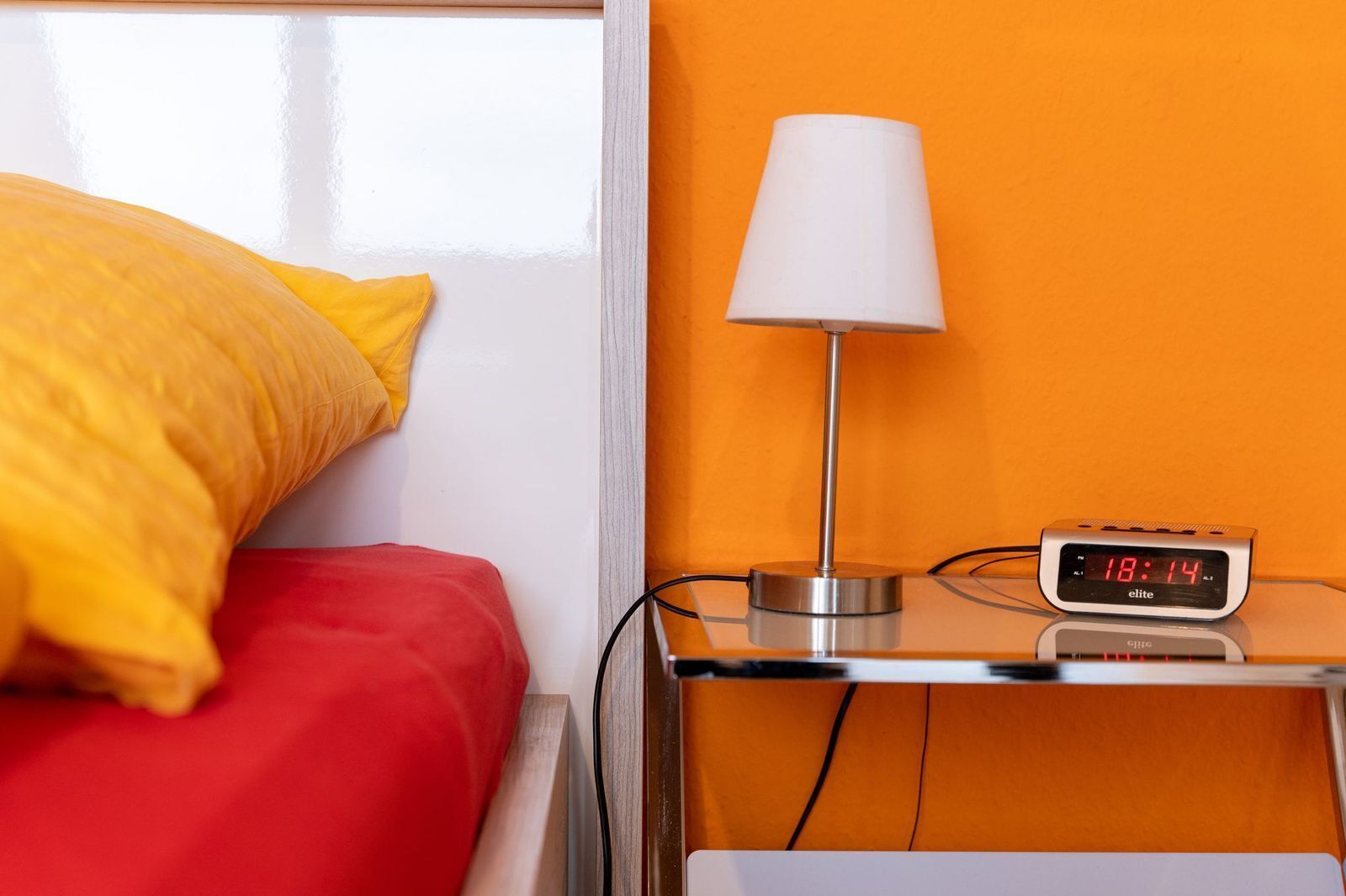 Ein Bett mit gelbem Kopfkissen und roter Decke, ein Nachttisch mit Lampe und digitaler Uhr, die 18:14 anzeigt, und eine orangefarbene Wand.