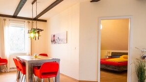 Un coin repas avec une table en bois et des chaises rouges, un tableau représentant un paysage urbain accroché au mur, et un lit recouvert d’une courtepointe rouge et jaune dans la pièce adjacente.