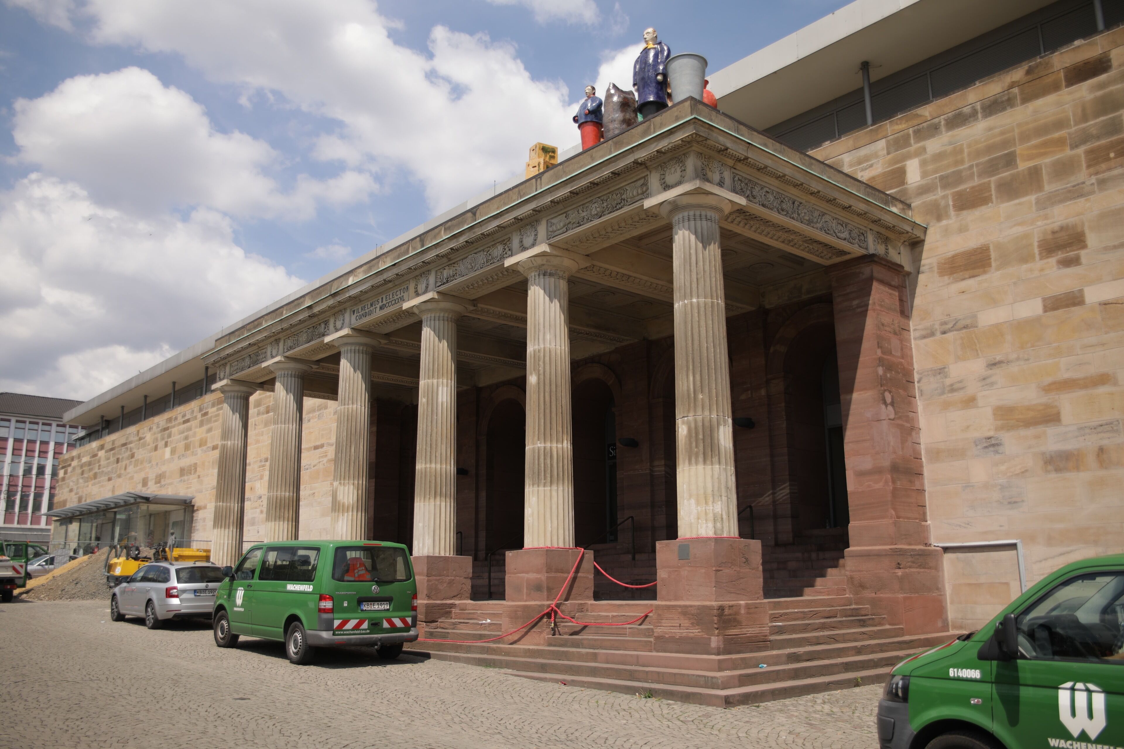 Un bâtiment à l’architecture classique, doté de colonnes et d’un fronton. Des véhicules sont stationnés devant.