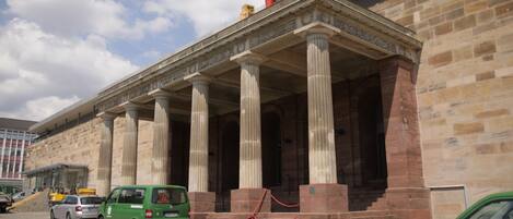 Un bâtiment à l’architecture classique, doté de colonnes et d’un fronton. Des véhicules sont stationnés devant.