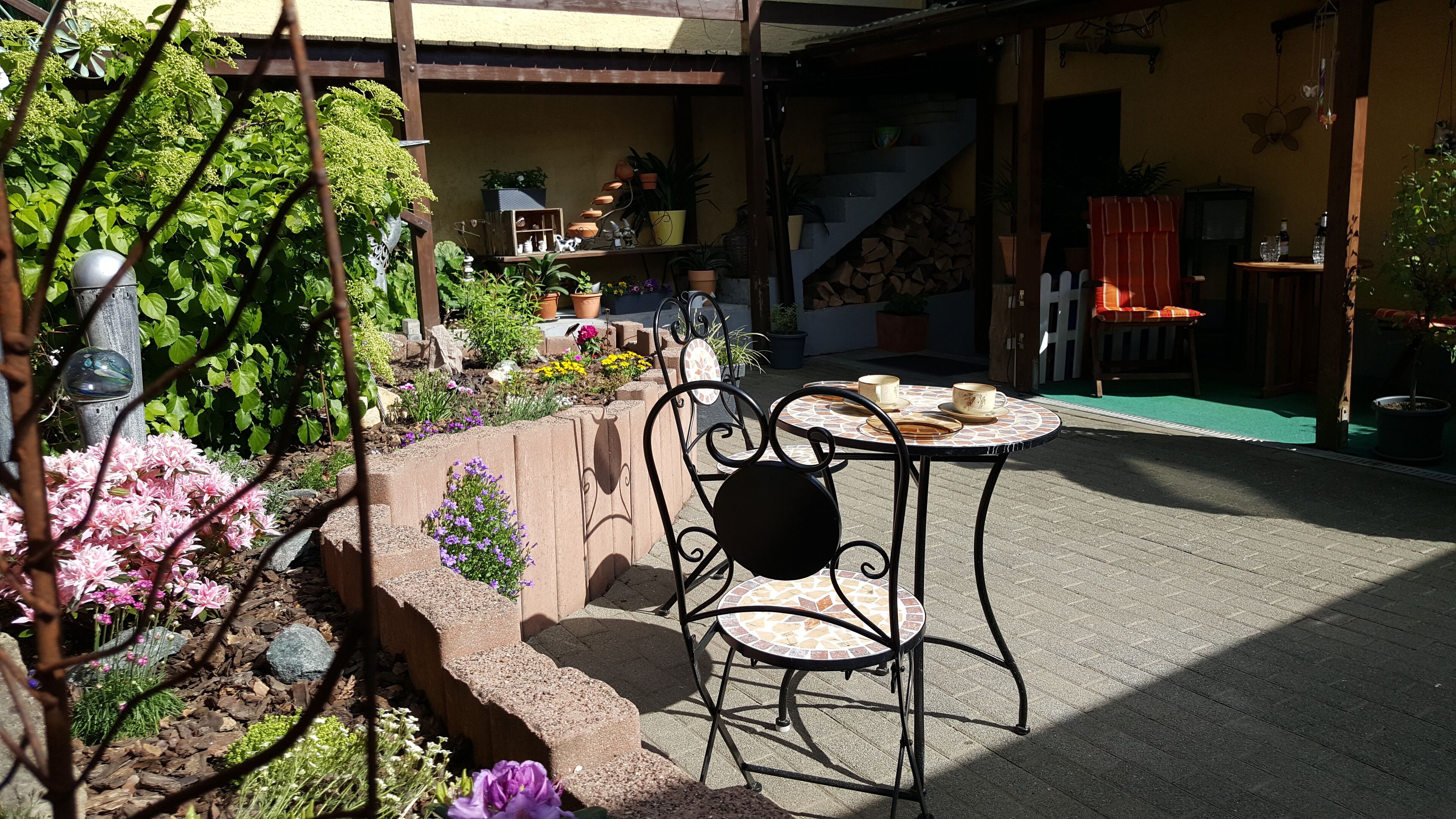 A patio area with a round table and chairs, surrounded by potted plants and flowers.