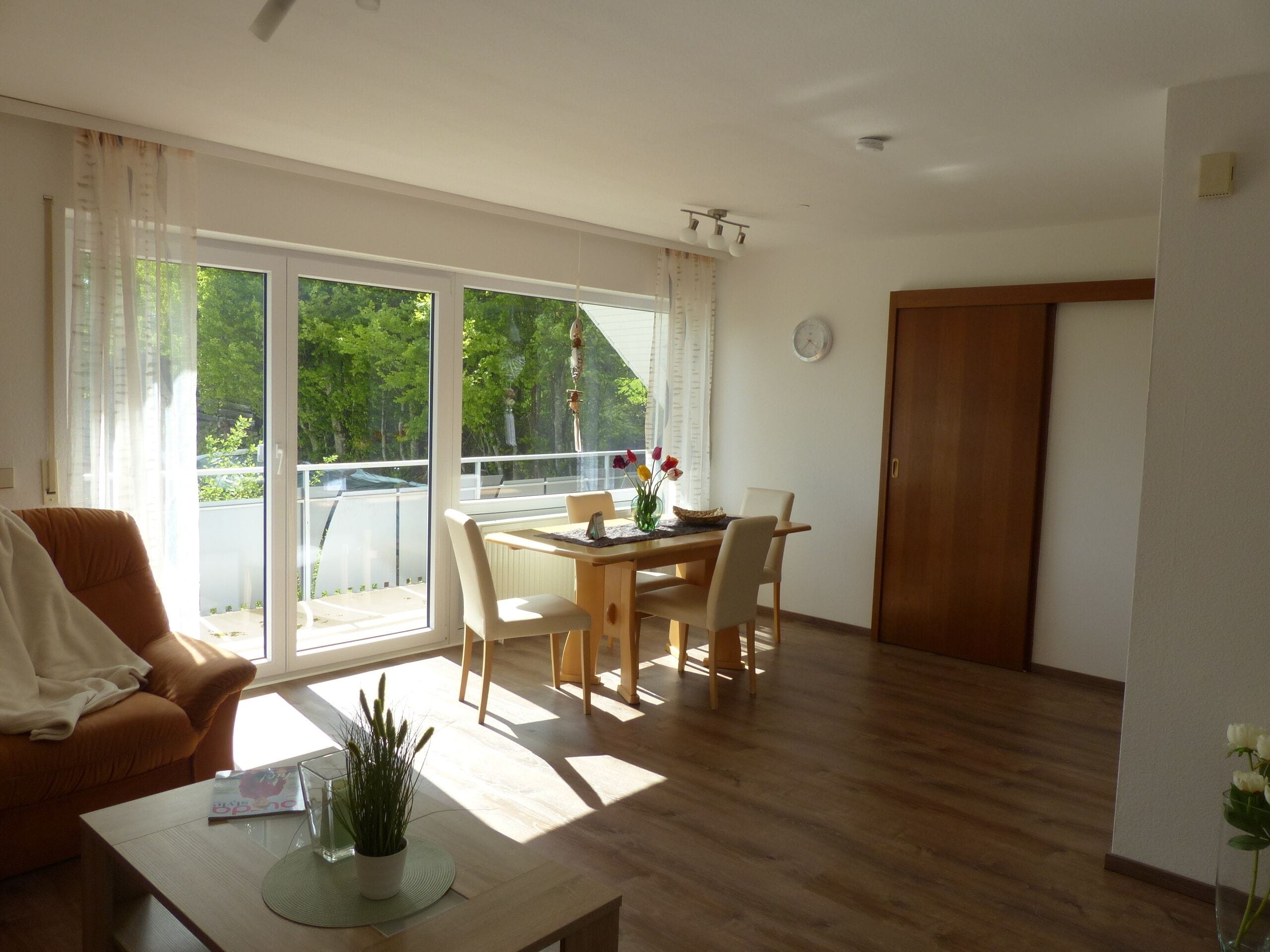 Ein geräumiges Wohnzimmer mit Holzboden, einem Esstisch mit Stühlen, einer Couch und einer großen Schiebeglasdoor, die auf einen Balkon führt.