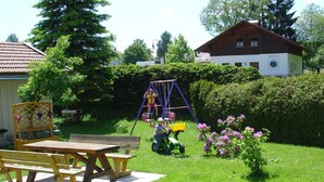 A wooden table and benches in a garden with a swing set and a lawnmower.