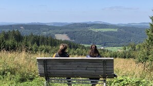 Deux personnes sont assises sur un banc, face à un paysage pittoresque composé de collines et d’arbres.