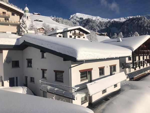 A snow-covered building with multiple balconies, a ski resort in the background, and a mountain range.