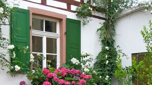Une terrasse avec une table et des chaises, entourée de fleurs roses et blanches en pleine floraison, et un bâtiment blanc avec des volets verts.