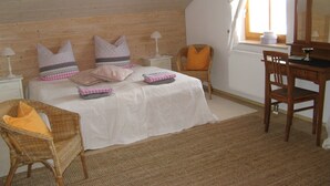 A bedroom with a sloped ceiling, a bed with multiple pillows, a wicker chair, a wooden desk, and a window with curtains. - Vacation home in a fantastic location in Upper Bavaria (Oberhausen)