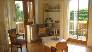 A room with a wooden floor, a table with chairs, a bookshelf, and a view of greenery outside. - Vacation home in a fantastic location in Upper Bavaria (Oberhausen)