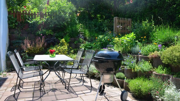 A patio with a table and chairs, a barbecue grill, and a lush garden with various plants and flowers. - Franconian idyll at Bamberg (Stegaurach)