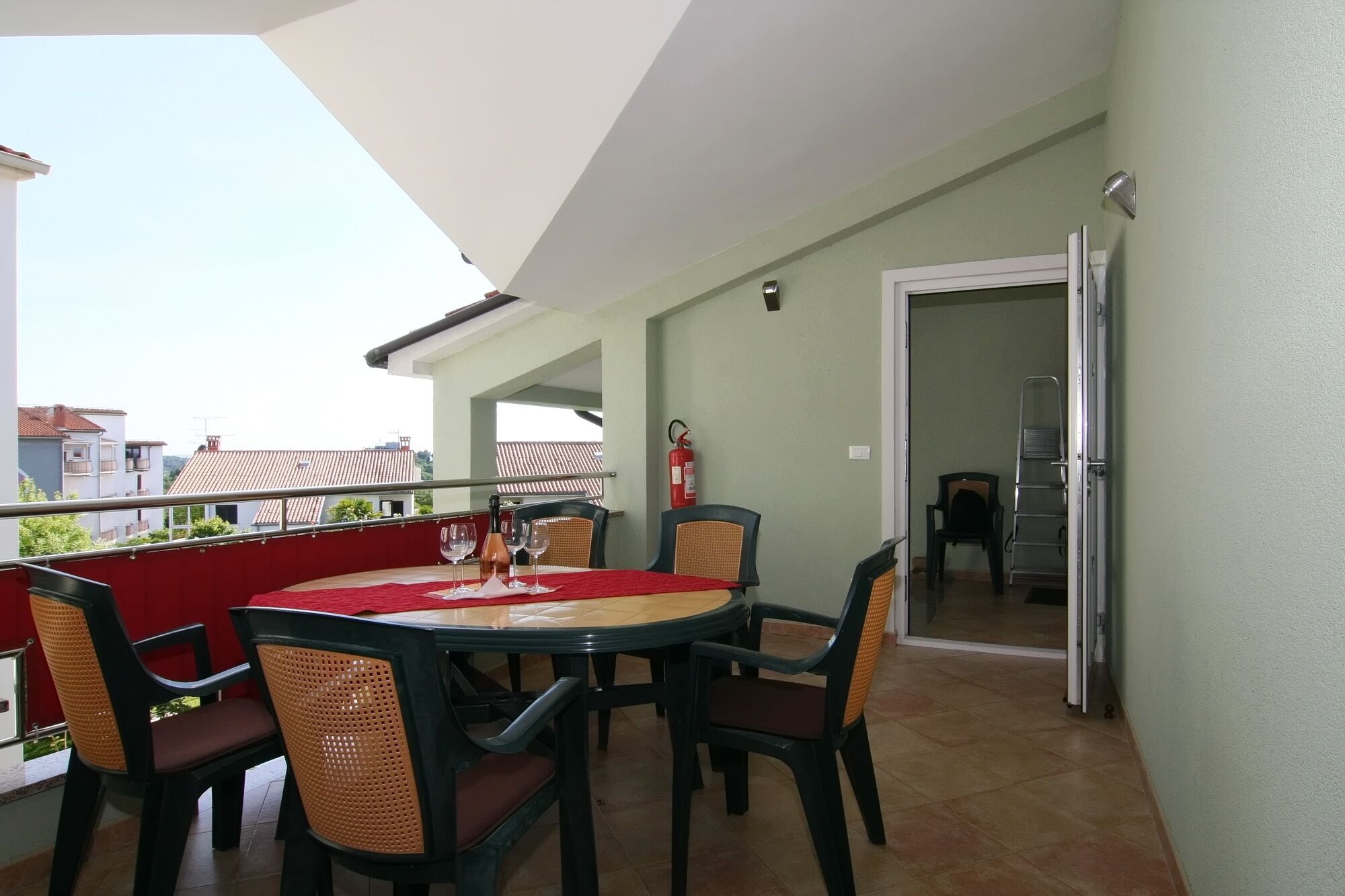 Un balcone con tavolo apparecchiato per quattro persone, sedie, un estintore e una porta che dà su un ambiente interno.